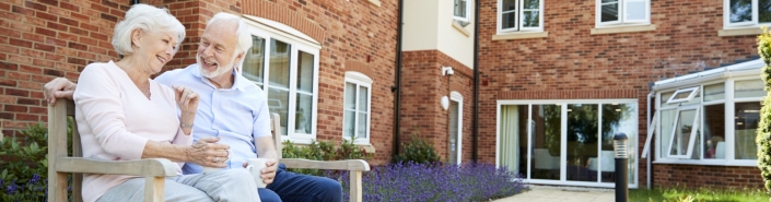 Retired Couple Sitting On Bench With Hot Drink In Assisted Living Facility